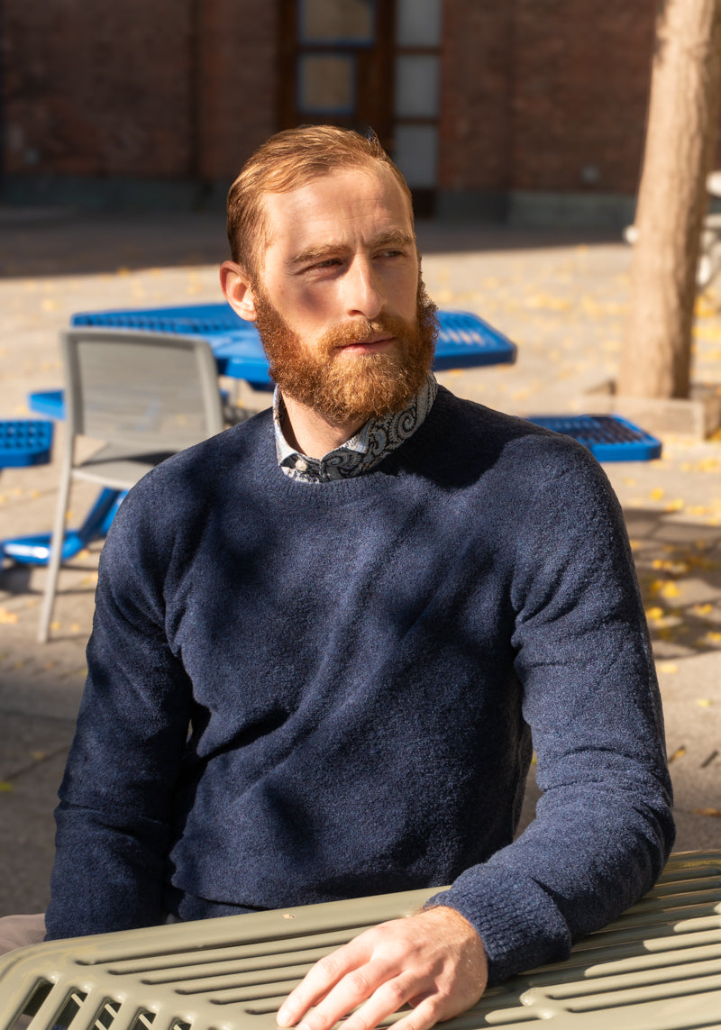 A man sitting outdoors wearing a blue boucle crewneck sweater.